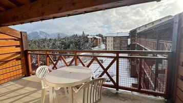 Un balcon en bois, agrémenté d’une table et de chaises, d’où l’on peut admirer un paysage montagneux enneigé et des bâtiments.