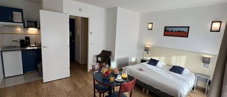 A hotel room with a bed, bedside tables, a desk with a chair, a small kitchenette, and a dining area with a table and chairs.