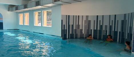 An indoor swimming pool with clear water, a tiled wall with vertical patterns, and a ceiling with air vents.