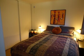A bedroom with a large bed, two bedside tables, a nightstand, a wall-mounted painting, and a sliding door. - Vechtdal Chalet de Eik (Rheezerveen)