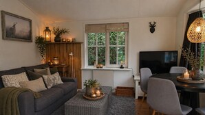 A cozy living room with a grey sofa, a small table with candles, a TV, and a window with a view of greenery. - Vechtdal Chalet de Berk (Rheezerveen)