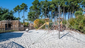 Een zandstrand met speeltoestellen, een houten constructie en een hoge boom.