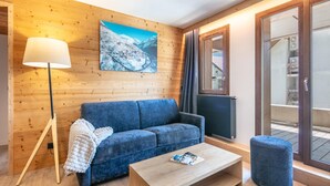 A modern living room with a blue sofa, a wooden floor, a standing lamp, a painting on the wall, and a view through a glass door. - 2-Person Studio near Ski Lifts (VAUJANY (ISERE))