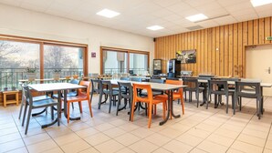 Uma área de refeições com mesas, cadeiras, uma máquina de vendas automática e grandes janelas.
