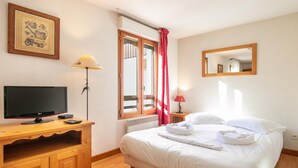 Quarto de hotel com uma cama, televisão sobre suporte de madeira, espelho e janela com cortinas.