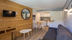 A modern interior with a wooden wall, a flat-screen TV, a round mirror, a dining area with chairs, a sofa, and a kitchen in the background. - Central Auris-en-Oisans Stay for 4 (Germ)