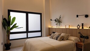 Ein Schlafzimmer mit großem Fenster, einem Nachttisch mit Lampe, einem Blumentopf und einem Bett mit beiger Decke.