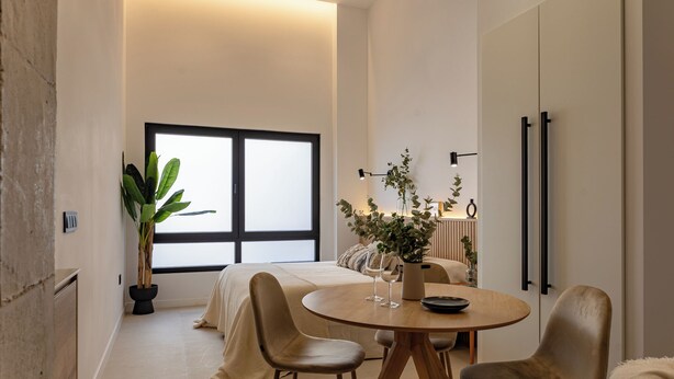 Ein modernes Schlafzimmer mit großem Fenster, einem Nachttisch mit Pflanze und einem runden Esstisch mit Stühlen.
