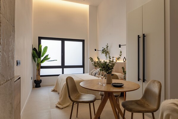 A modern bedroom with a large window, a bedside table with a plant, and a round dining table with chairs. - Almirante Suites B (València)