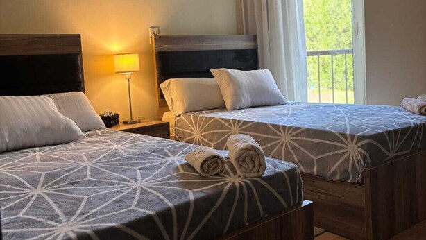 A hotel room with two beds, a nightstand, a lamp, and a window with curtains. - Luxury Stay in Sheikh Zayed -Private Garden + Swimming Pool (6th of October City)