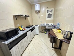 Cozinha compacta com micro-ondas, torradeira e pia. Uma prateleira pequena acomoda xícaras e chaleira.