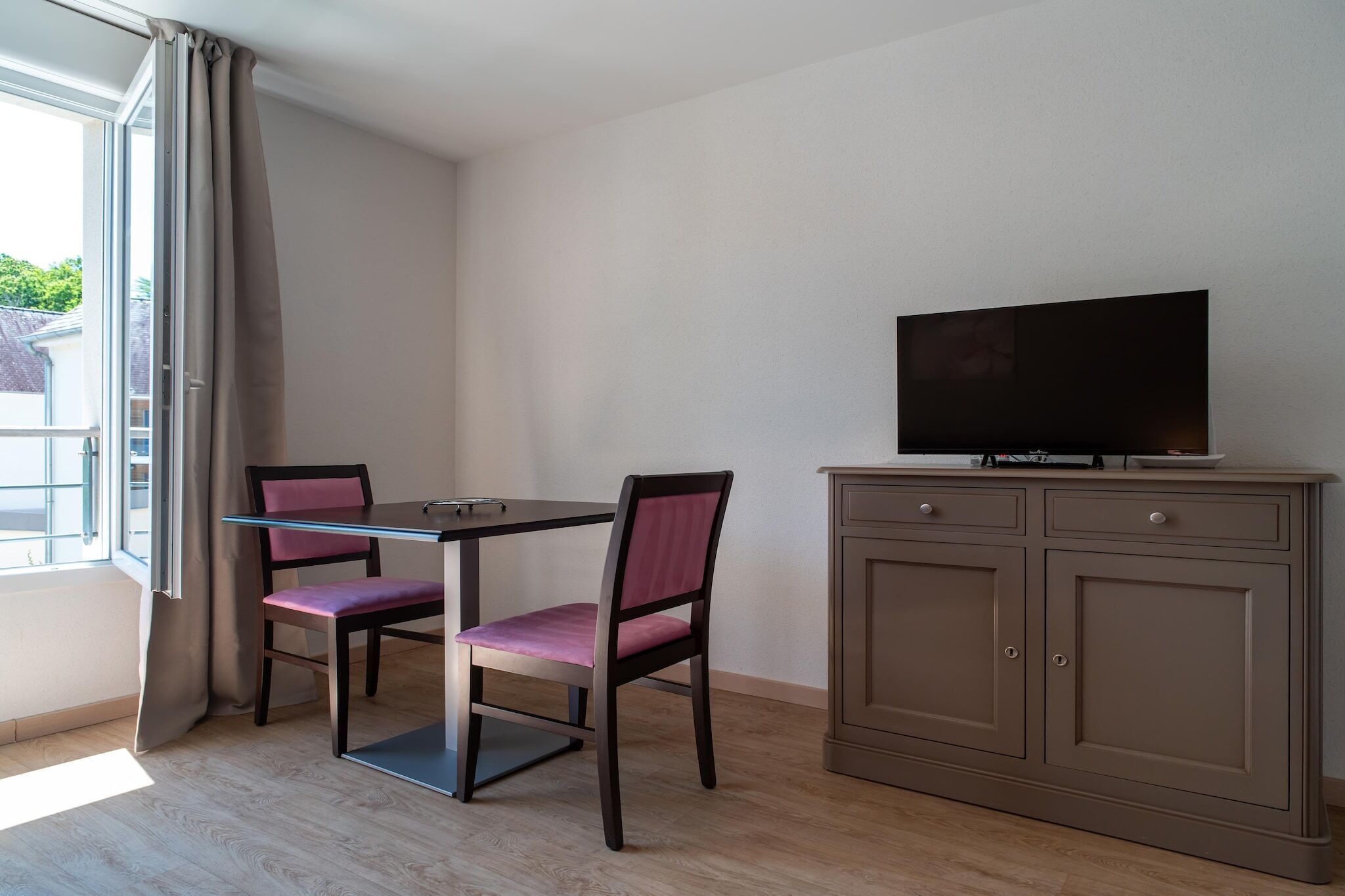 Quarto com uma mesa, duas cadeiras, televisão em um armário e uma janela com cortinas.