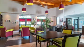 Ein modernes Interieur mit Holzbalkendecke, einem zentralen Esstisch, bunten Stühlen und Topfpflanzen.