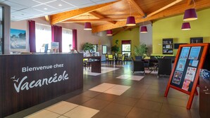 Un espace d’accueil avec un comptoir de réception en bois, des suspensions violettes, un tableau d’affichage et un coin salon avec des tables et des chaises.