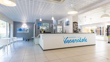 A reception desk with a sign reading 'BIENVENUE CHEZ Vacanceole,' a white interior with tiled floors, pendant lighting, and a view of the outside.