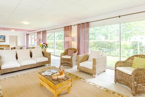 A spacious living area with wicker furniture, large windows, and a view of greenery.