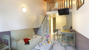 Ein möbliertes Zimmer mit einer Couch, einem Tisch mit Früchten und einem Glas, einem Stuhl, einer Treppe und einem an der Wand befestigten Fernseher.