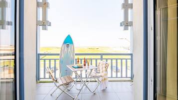 Un balcon avec une planche de surf, deux chaises, une table avec des bouteilles et une vue sur la plage.