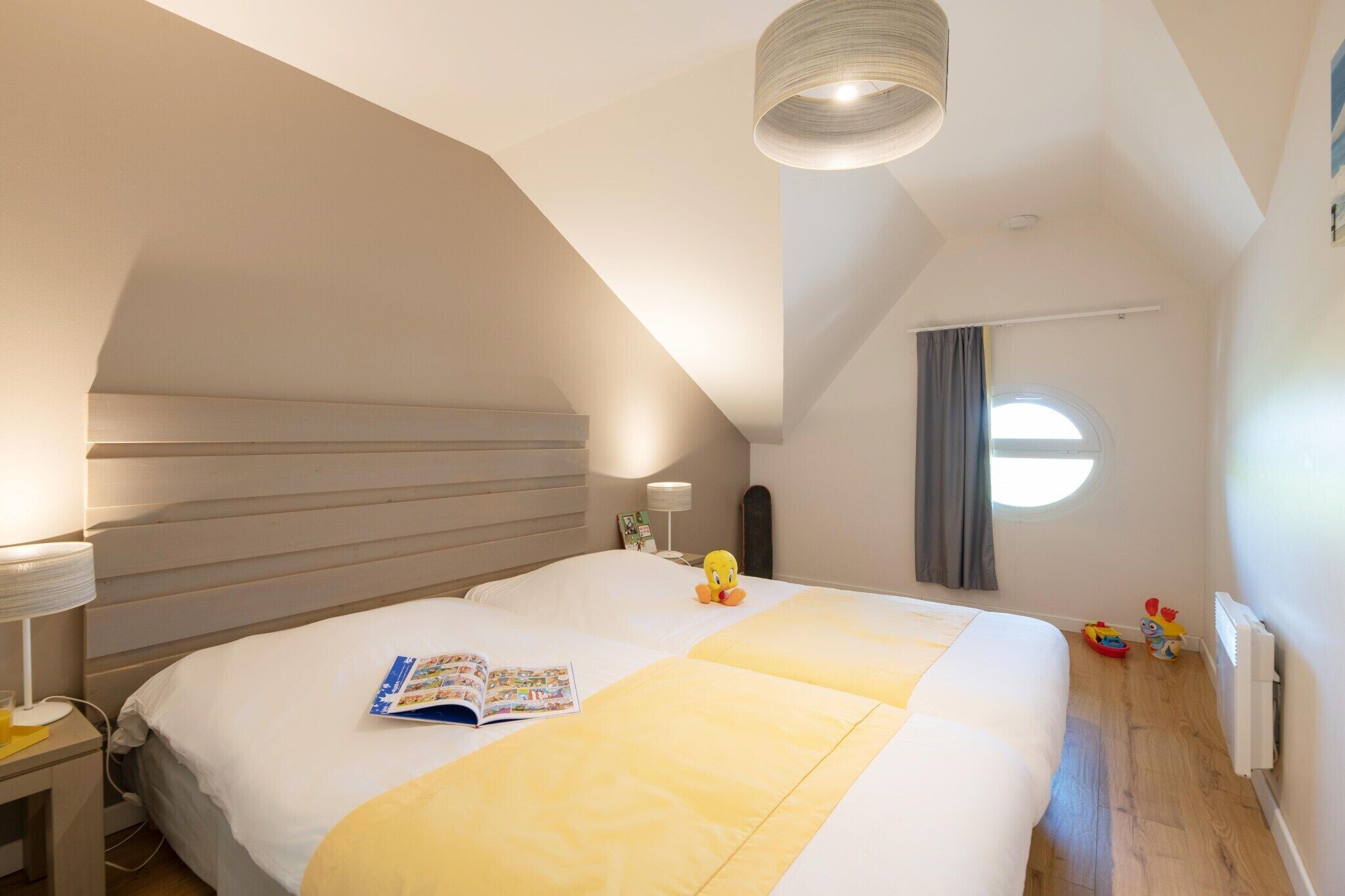 Une chambre à coucher avec un grand lit, une table de chevet avec une lampe, une fenêtre avec des rideaux et un luminaire au plafond.
