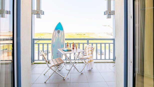 Ein Balkon mit einem Surfbrett, zwei Stühlen, einem Tisch mit Flaschen und Blick auf den Strand.