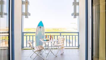 Un balcon avec une planche de surf, deux chaises, une table avec des bouteilles et une vue sur la plage.