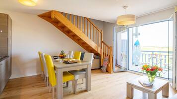 Un coin repas avec un escalier en bois, une table à manger avec des chaises, une petite table sur laquelle est posé un vase, et une vue à travers une porte vitrée.