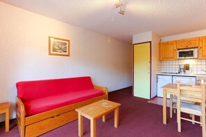 Un espace de vie compact comprenant un canapé rouge, une table basse en bois et une kitchenette dotée de meubles de rangement en bois.