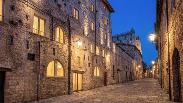 A historic stone building with arched windows and a tower, a cobblestone street, and street lamps. - Edward Historic House (Gubbio)