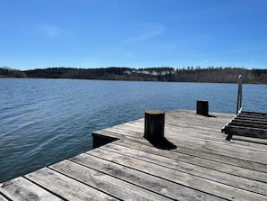 Un muelle de madera se adentra en un lago tranquilo, con una orilla boscosa al fondo.