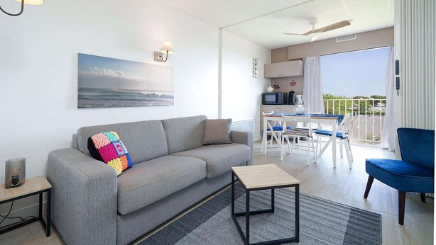 Ein modernes Wohnzimmer mit einem grauen Sofa, einem farbenfrohen Sitzkissen, einem kleinen Tisch und Blick auf den Balkon.