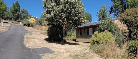 Un camino rural con una casa a la derecha, rodeado de árboles y hierba seca.
