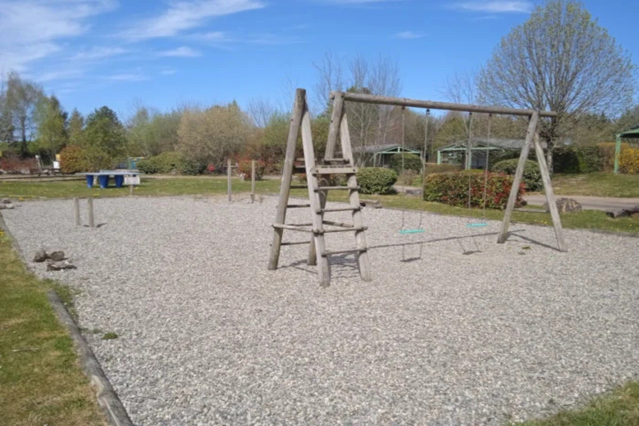 Une aire de jeux avec une balançoire en bois et une structure d’escalade sur une surface recouverte de gravier.