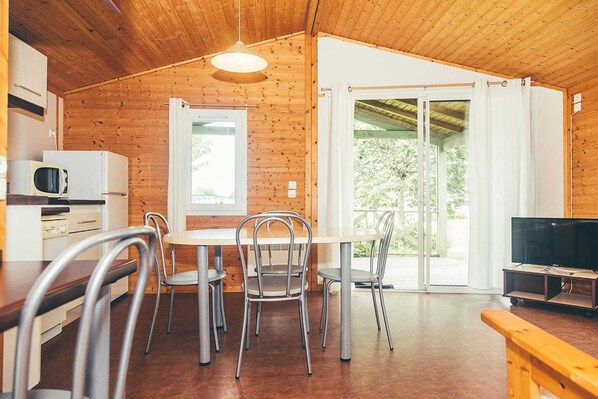 Un intérieur en bois comprenant une table à manger, des chaises, un téléviseur et une porte-fenêtre coulissante donnant sur un espace extérieur.