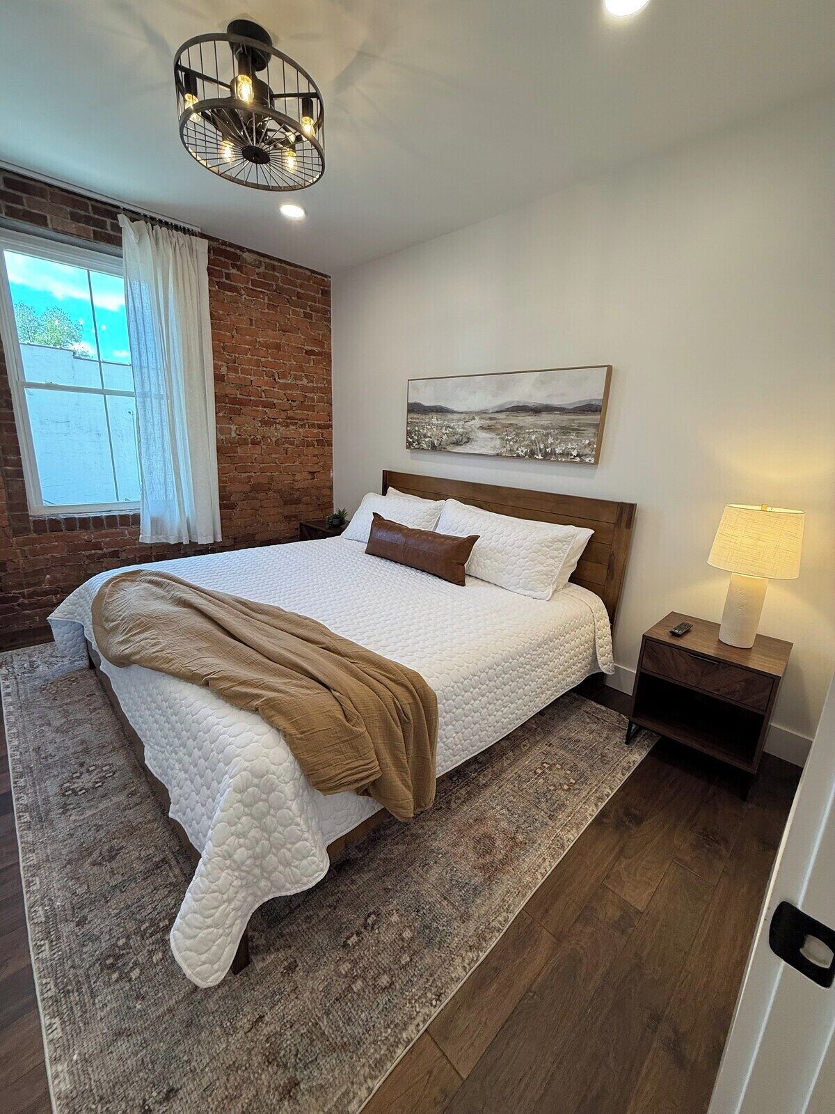 Quarto com cama, mesa de cabeceira, luminária, janela com cortinas, parede de tijolos e luminária no teto.