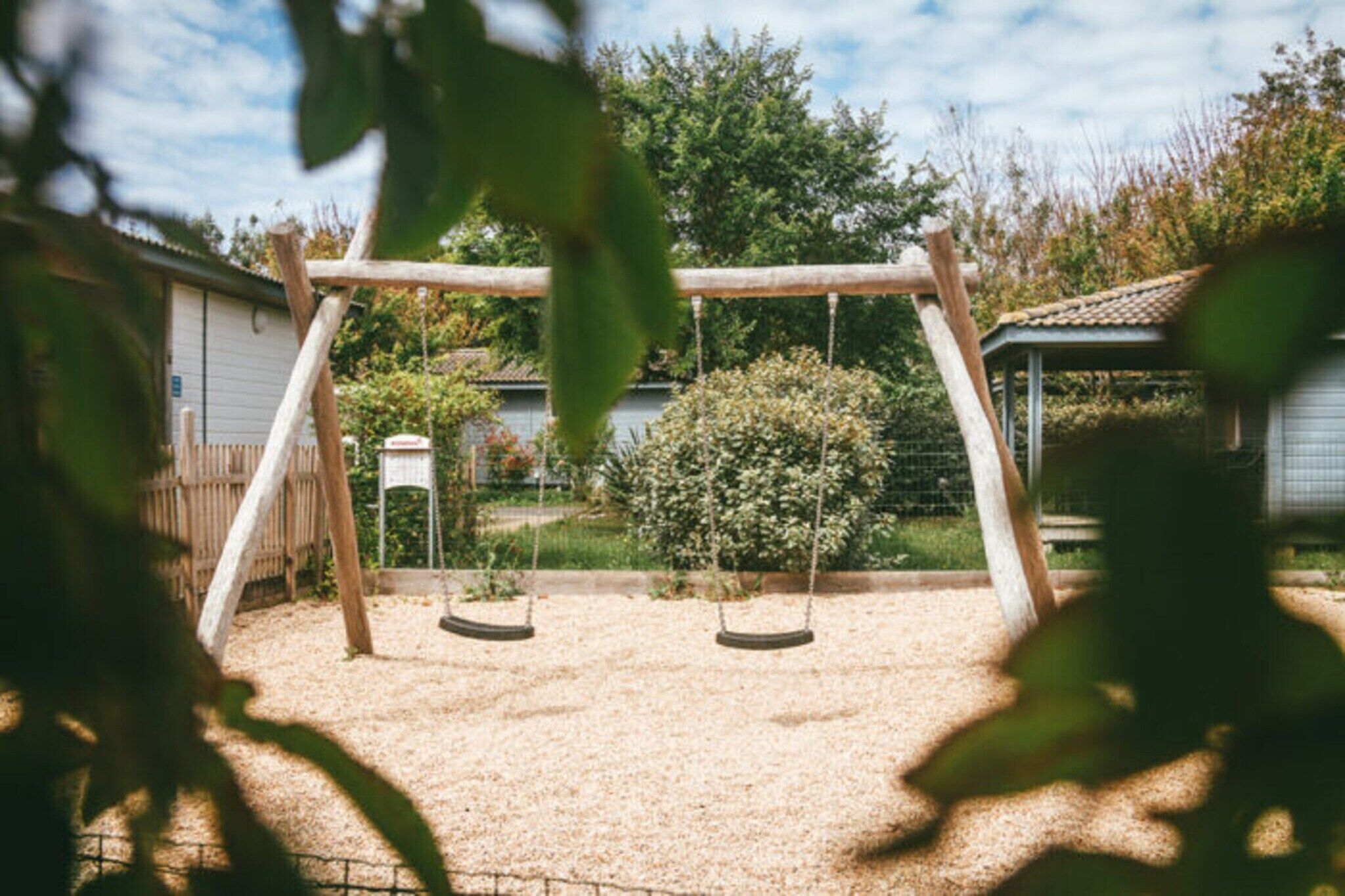 Ein hölzernes Schaukelgerüst in einem Garten mit Zaun, Strauch und Gartenhaus im Hintergrund.