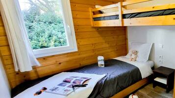 Une petite pièce en bois, aménagée comme une cabine, avec des lits superposés, une fenêtre donnant sur la verdure, une table de chevet avec un ours en peluche et un lit avec une couverture et un magazine.