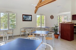 A dining area with tables and chairs, large windows, a wooden ceiling, and a potted plant.