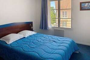 A bed with a blue quilt and white pillows, a wooden headboard, a window with a view of buildings, and a framed picture on the wall.