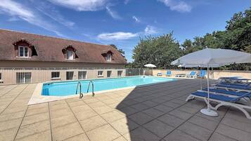 A swimming pool with lounge chairs and umbrellas, surrounded by a building with a tiled roof and a fence.