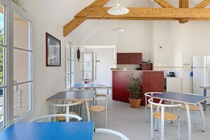 A kitchen with wooden beams, a dining area with tables and chairs, and a view through a window.