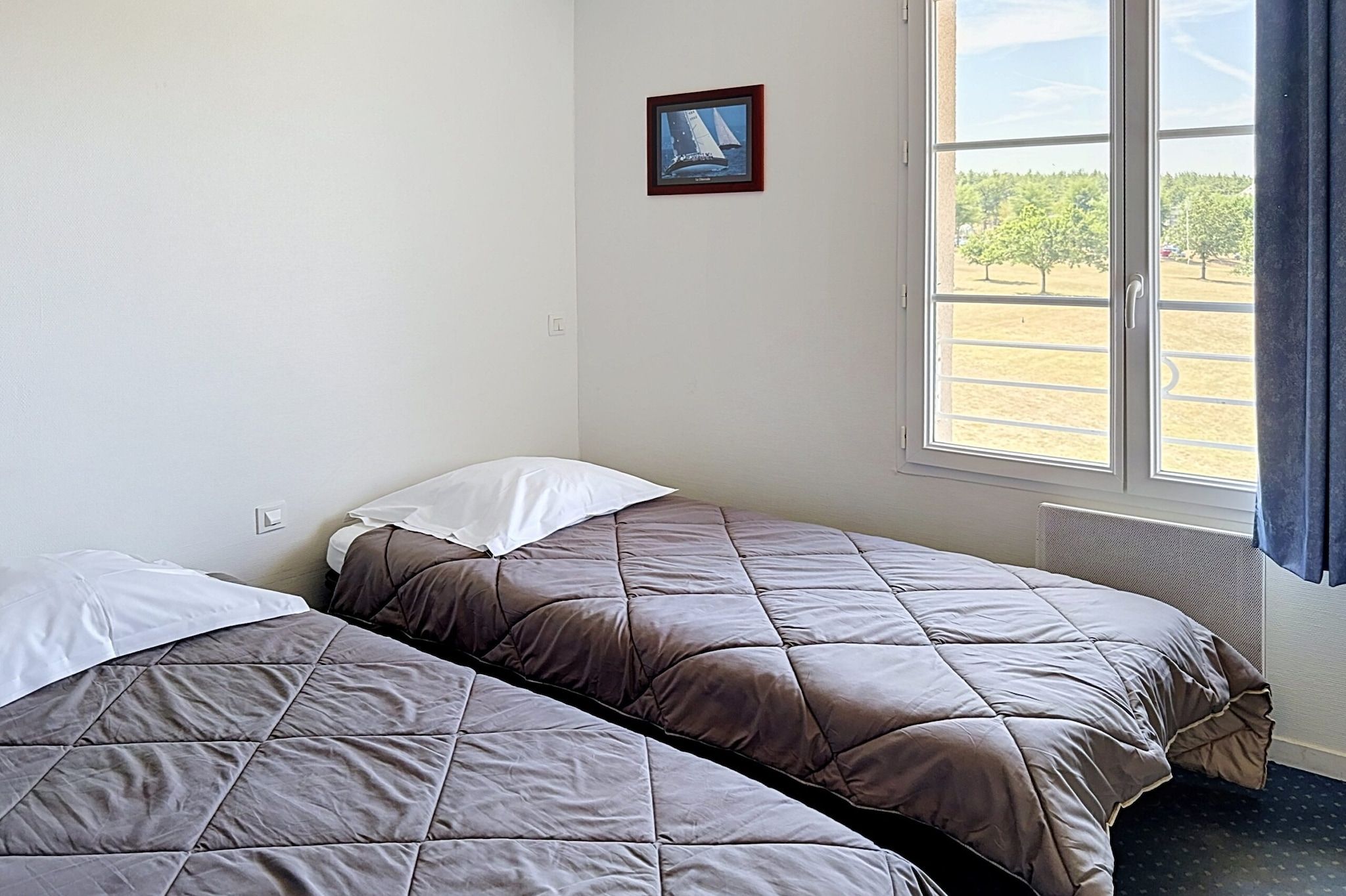 A room with two beds, a window with a view of trees, and a framed picture on the wall.