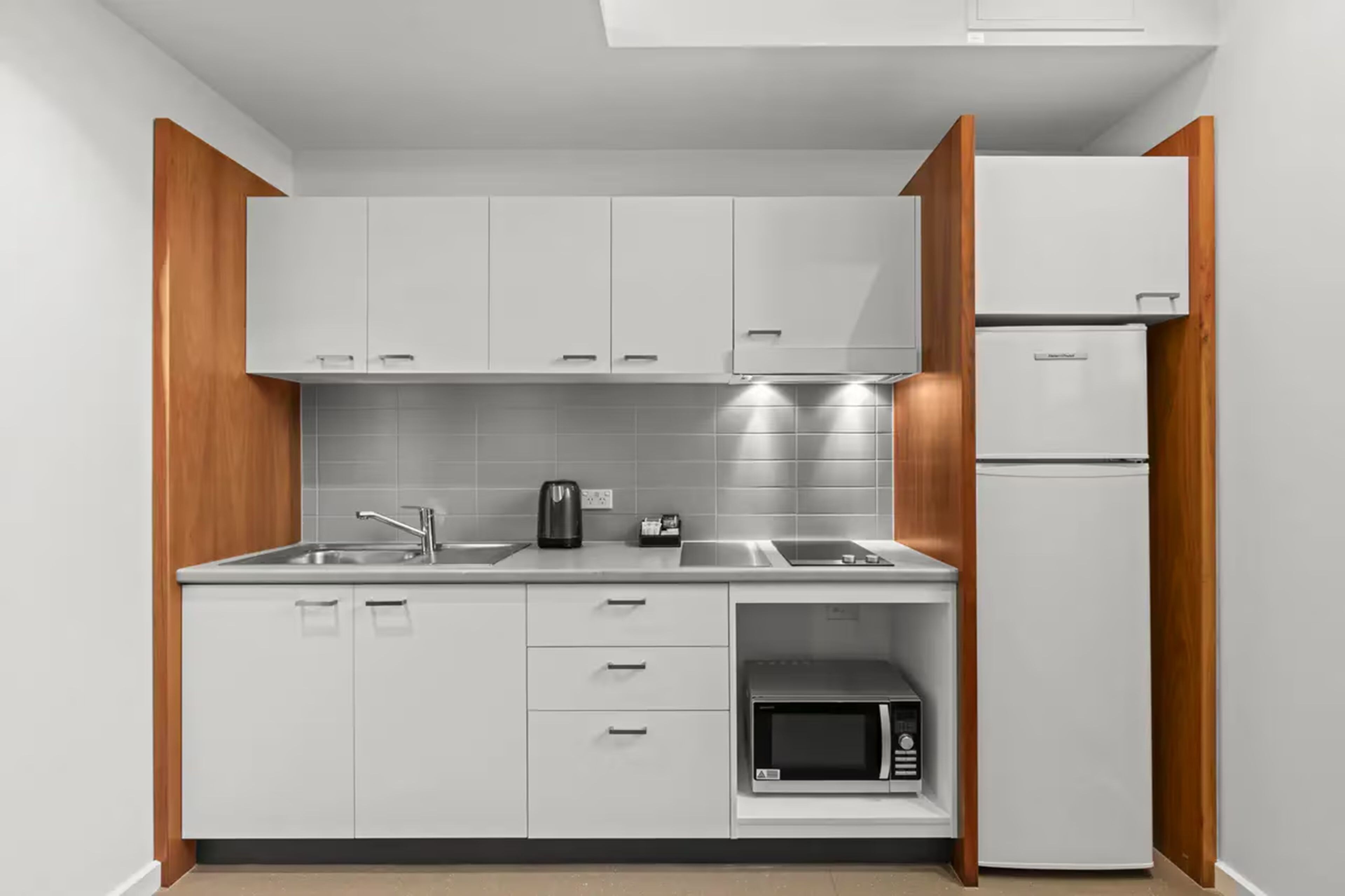 A compact kitchen with white cabinets, a stainless steel sink, a kettle, a microwave, and a refrigerator.