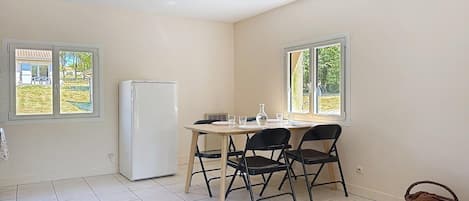 Une pièce comprenant une table à manger, des chaises, un réfrigérateur et des fenêtres donnant sur un jardin.