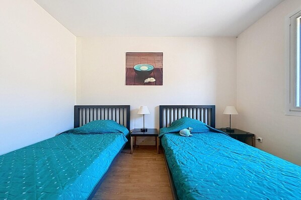 Habitación con dos camas individuales, cada una con una mesita de noche y una lámpara. Se observa una ventana a la derecha y un cuadro en la pared, sobre las camas.