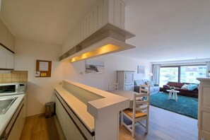 A compact kitchen with modern appliances, a dining area with a white table and chairs, and a living space with a sofa and a view through a large window. - Central Coastal Apartment for 4 (OOSTDUINKERKE)