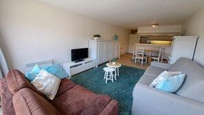 Ein Wohnzimmer mit einem braunen Sofa, einem grauen Sofa, einem Fernseher, einem kleinen Tisch mit einer Pflanze und einem Küchenbereich im Hintergrund.
