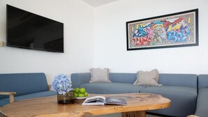 Ein modernes Wohnzimmer mit einem Flachbildfernseher, einem blauen Sofa, einem Holztisch mit Buch und Obstschale sowie einer farbenfrohen Graffiti-Kunst an der Wand.