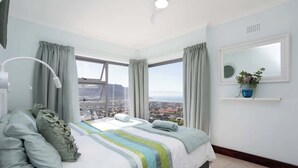Ein Schlafzimmer mit einem großen Bett, einer Nachttischlampe, einem Fenster mit Vorhängen, einem Spiegel und Blick auf eine Stadtlandschaft.