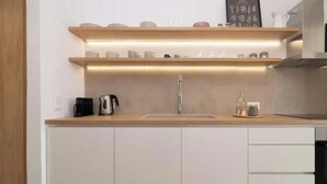 A modern kitchen with a built-in sink, stainless steel faucet, and a coffee maker. There are wooden shelves above the sink with various kitchen items. - Casa Tu Zante ❤️- center & sea view (for 6 guests) (Zakynthos)