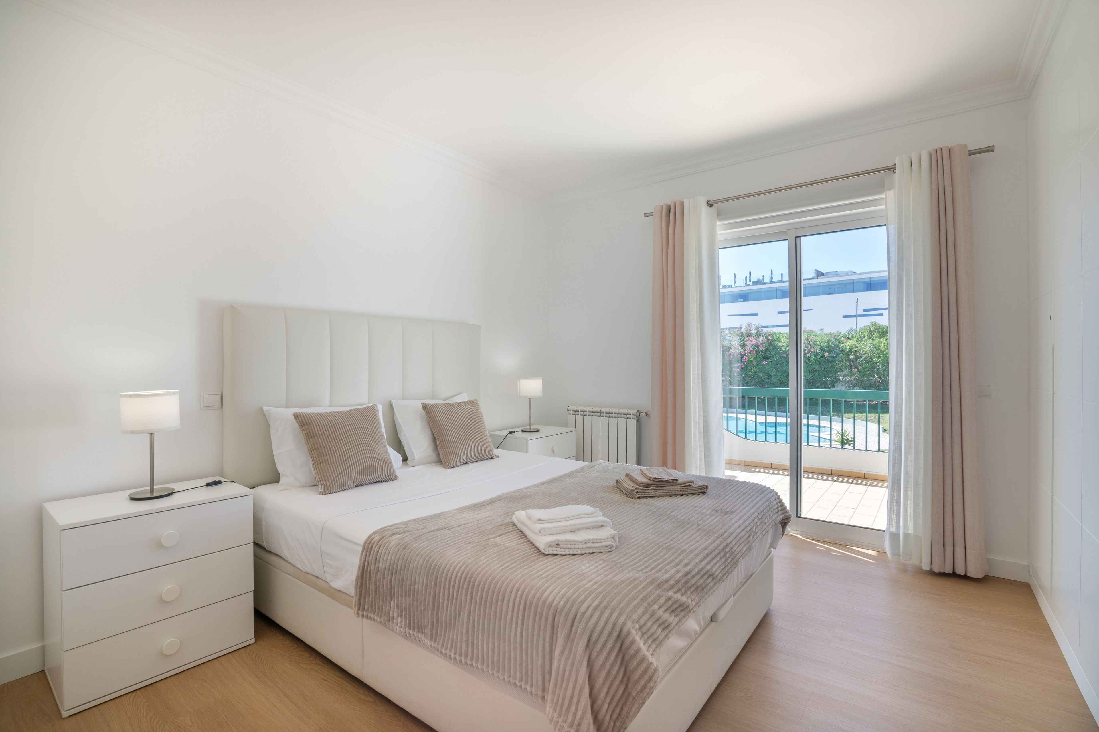 Een slaapkamer met een bed, nachtkastjes, een lamp, uitzicht op een zwembad en een balkon.
